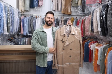 Photo of Dry-cleaning service. Happy man holding hanger with coat in plastic bag indoors