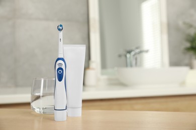 Photo of Electric toothbrush, tube with paste and glass of water on wooden table in bathroom. Space for text
