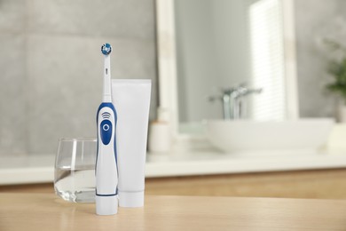 Electric toothbrush, tube with paste and glass of water on wooden table in bathroom. Space for text