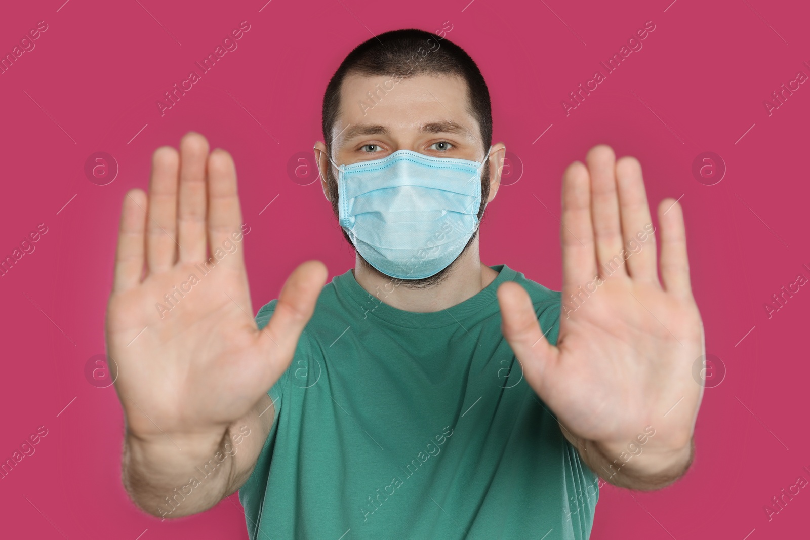 Photo of Man in protective mask showing stop gesture on pink background. Prevent spreading of coronavirus