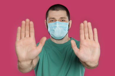 Photo of Man in protective mask showing stop gesture on pink background. Prevent spreading of coronavirus
