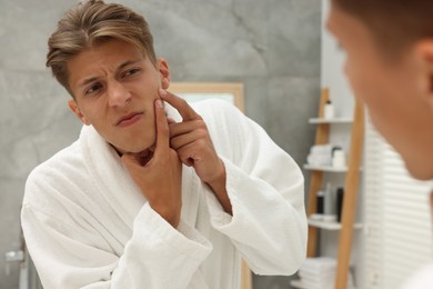 Photo of Upset young man looking at mirror and popping pimple on his face indoors. Acne problem