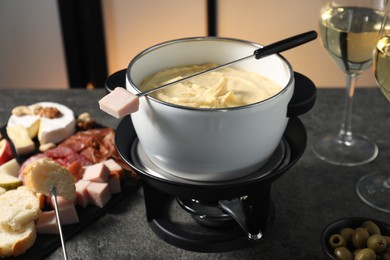 Forks with pieces of ham, bread and fondue pot with melted cheese on grey table, closeup