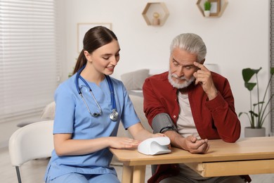 Young healthcare worker measuring senior man's blood pressure at wooden table indoors