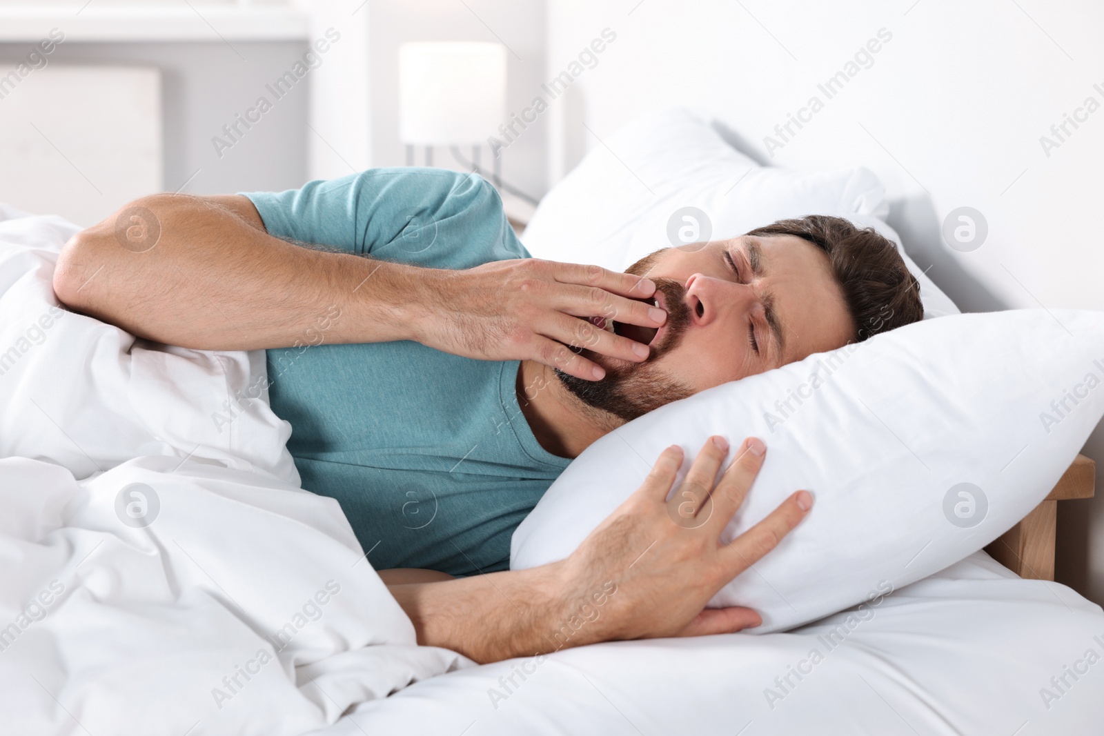 Photo of Handsome man yawning while lying on pillow in bed at home