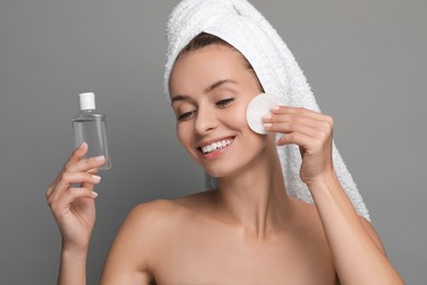 Photo of Smiling woman removing makeup with cotton pad and holding bottle on grey background