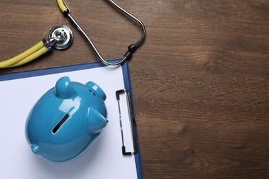 Light blue ceramic piggy bank, clipboard and stethoscope on wooden table, flat lay with space for text. Medical insurance