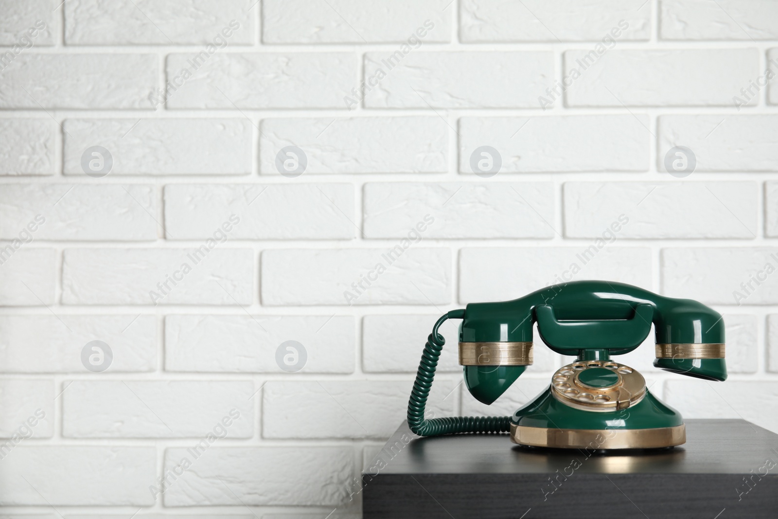 Photo of Green vintage corded phone on small black table near white brick wall. Space for text