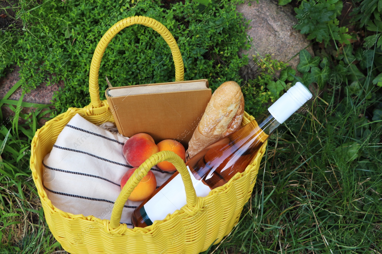 Photo of Yellow wicker bag with book, peaches, baguette and wine on green grass outdoors, above view