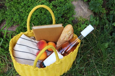 Photo of Yellow wicker bag with book, peaches, baguette and wine on green grass outdoors, above view