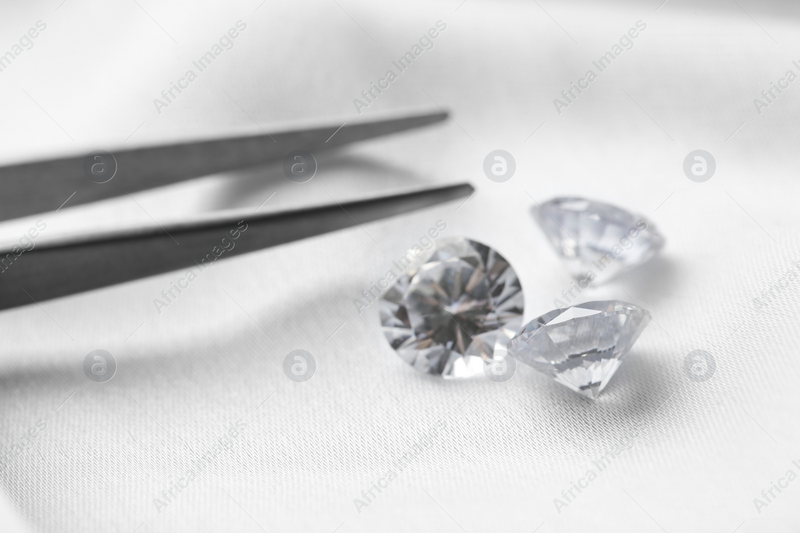 Photo of Beautiful shiny diamonds and tweezers on white fabric, closeup