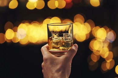Man holding glass of whiskey with ice cubes against blurred lights, closeup.