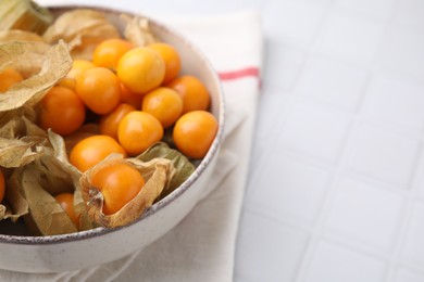 Photo of Ripe physalis fruits with calyxes in bowl on white tiled table, closeup. Space for text