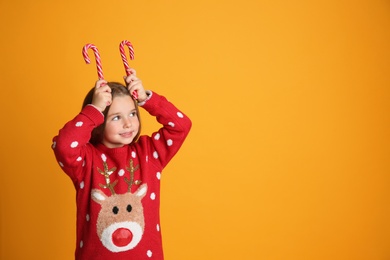 Cute little girl in Christmas sweater holding candy canes on yellow background, space for text