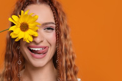 Photo of Beautiful young hippie woman covering eye with sunflower and showing her tongue on orange background, closeup. Space for text