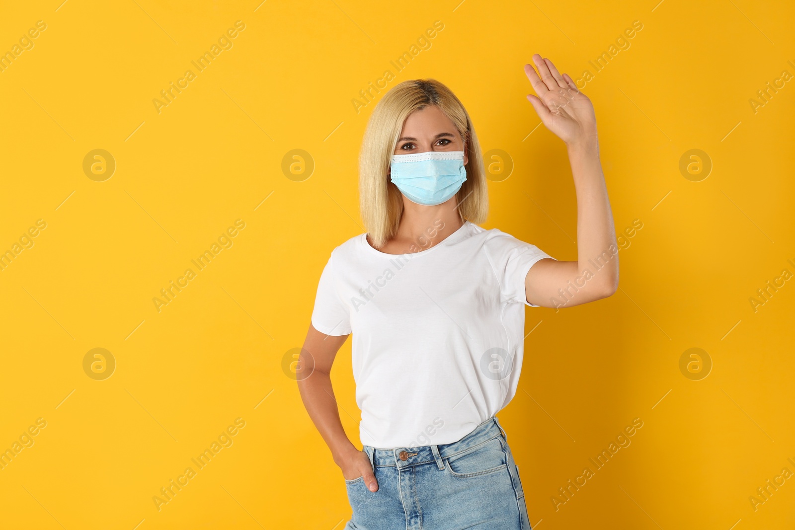 Photo of Woman in protective mask showing hello gesture on yellow background. Keeping social distance during coronavirus pandemic