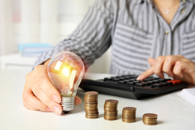 Photo of Woman with light bulb, calculator and coins at white table, closeup. Power saving