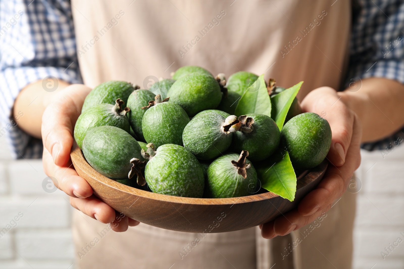 Photo of Woman holding fresh green feijoa fruits in bowl against white brick wall, closeup