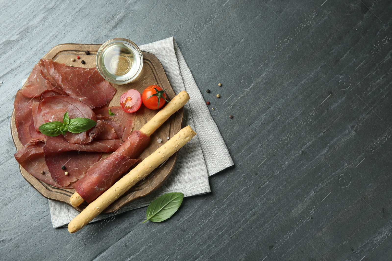 Photo of Delicious bresaola, tomato, grissini sticks and basil leaves on grey textured table, top view. Space for text