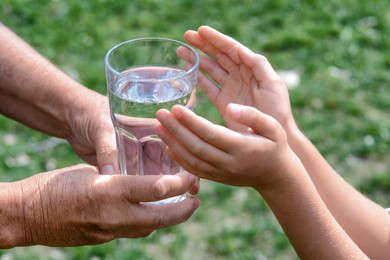 Photo of Child giving glass of water to elderly woman outdoors, closeup