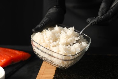 Photo of Chef in gloves taking cooked rice for sushi with spoon at dark table, closeup