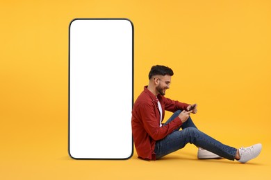 Image of Man with mobile phone sitting near huge device with empty screen on orange background. Mockup for design