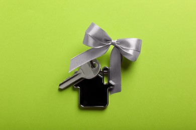 Key with trinket in shape of house and bow on light green background, top view. Housewarming party