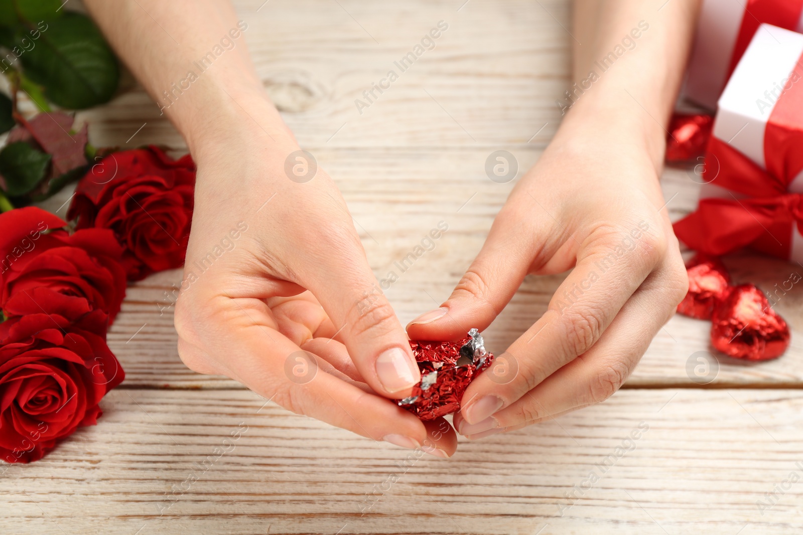 Photo of Woman unwrapping heart shaped chocolate candy at white wooden table, closeup