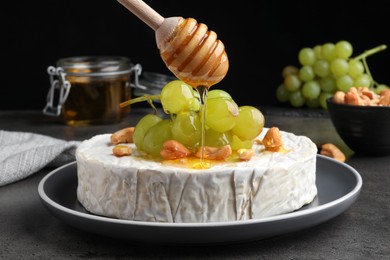 Photo of Pouring honey onto brie cheese served with grape and cashew nuts on dark grey table, closeup