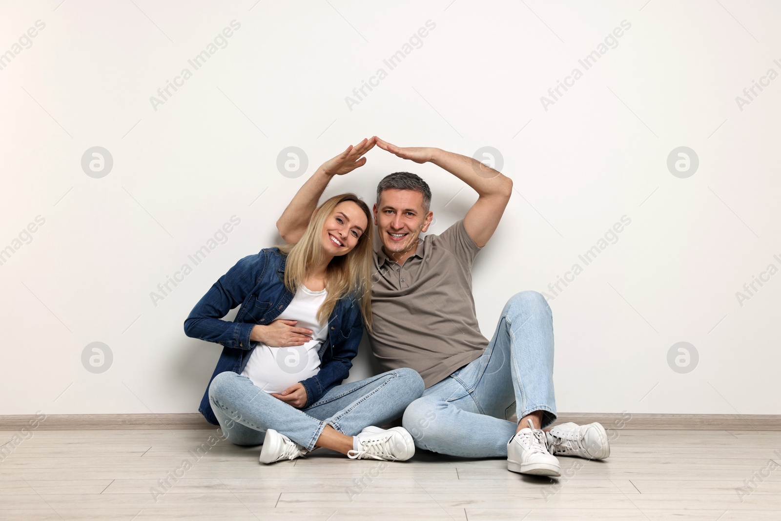 Photo of Young family housing concept. Pregnant woman with her husband forming roof with their hands while sitting on floor indoors