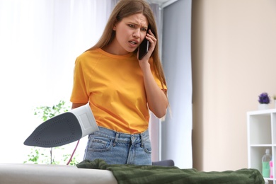 Photo of Emotional woman talking on phone while ironing clothes at home. Space for text