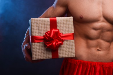 Young man with muscular body holding Christmas gift box on color background, closeup