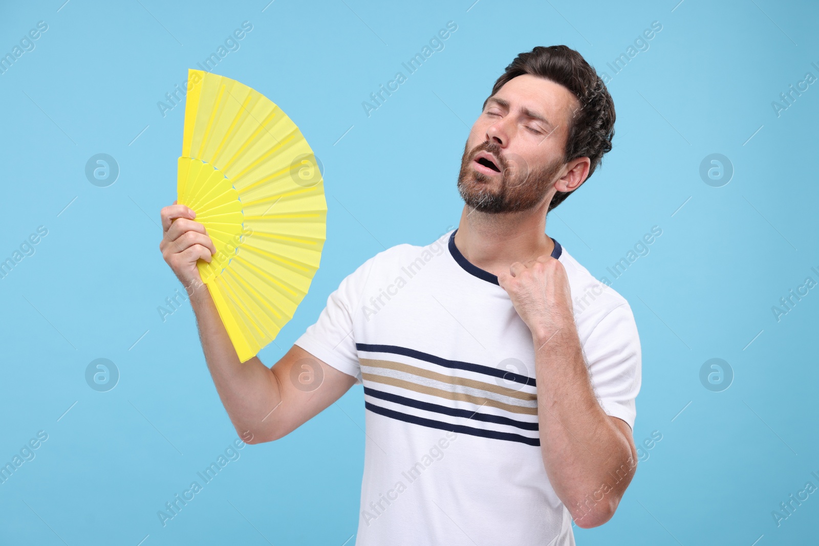 Photo of Unhappy man with hand fan suffering from heat on light blue background