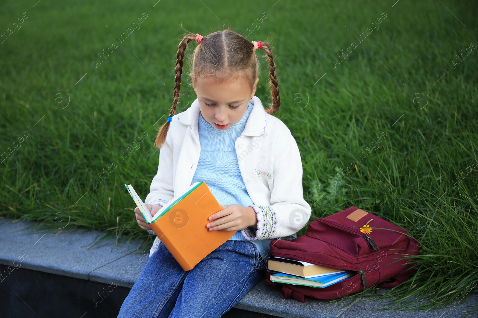 Photo of Cute little girl with backpack reading textbook on city street