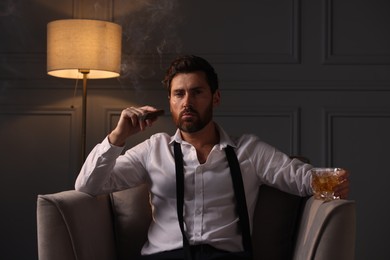 Photo of Handsome man with glass of whiskey smoking cigar in armchair at home