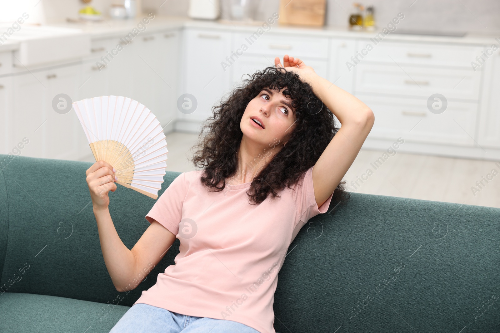 Photo of Young woman waving hand fan to cool herself on sofa at home