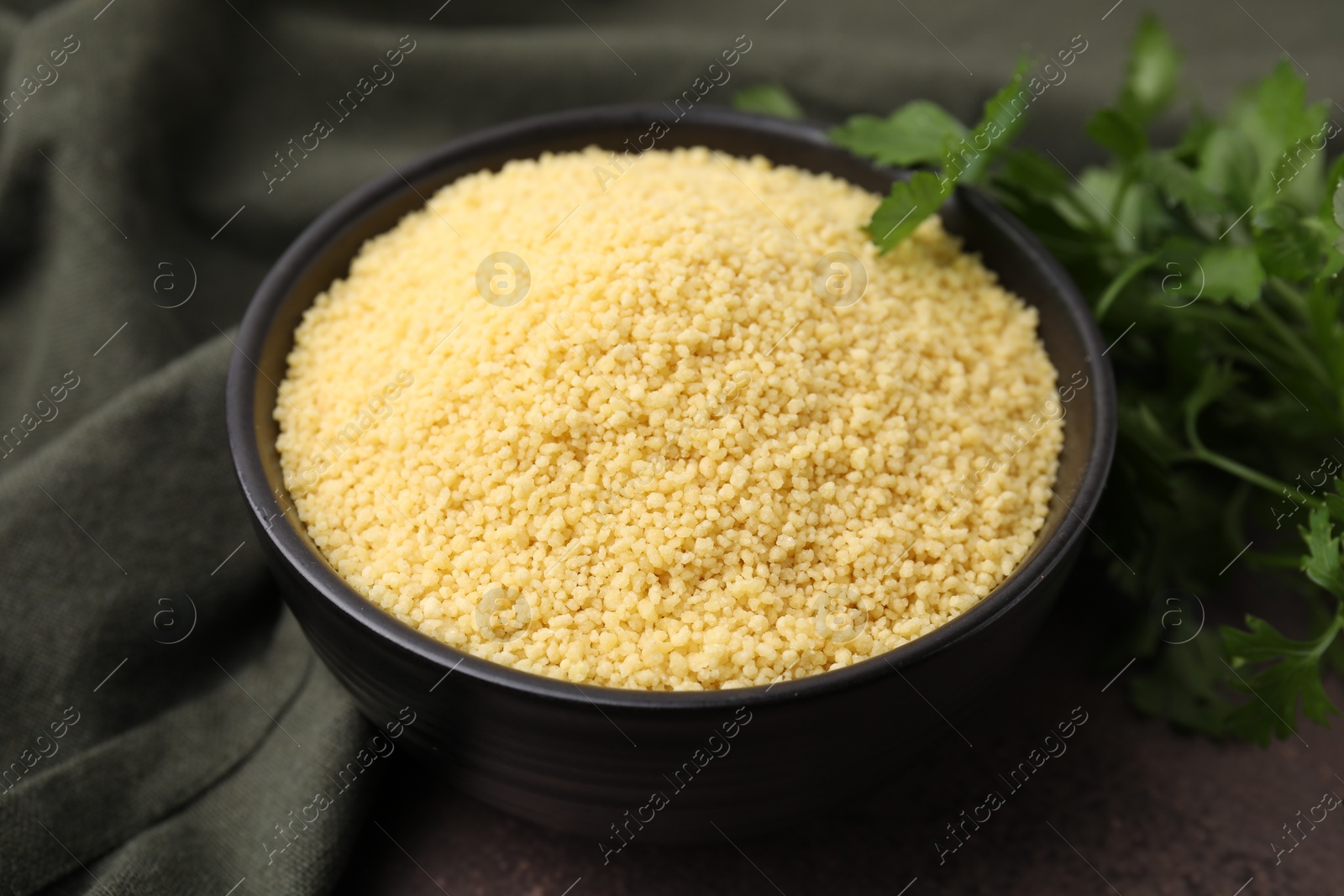 Photo of Raw couscous in bowl and parsley on table, closeup