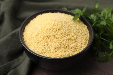 Photo of Raw couscous in bowl and parsley on table, closeup