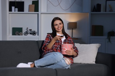 Photo of Happy woman holding popcorn bucket and changing TV channels with remote control at home in evening
