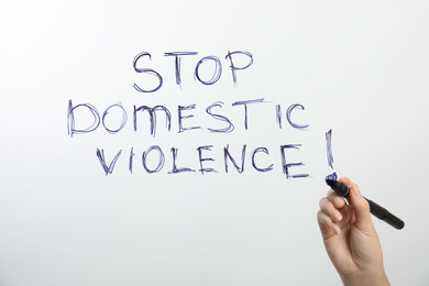 Photo of Woman writing phrase STOP DOMESTIC VIOLENCE on glass against white background, closeup