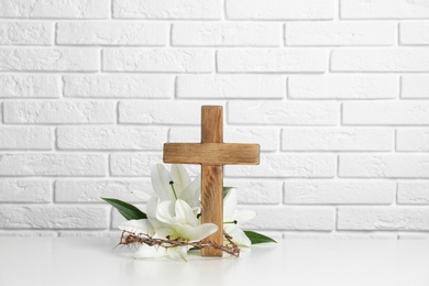 Photo of Wooden cross, crown of thorns and blossom lilies on table against brick wall