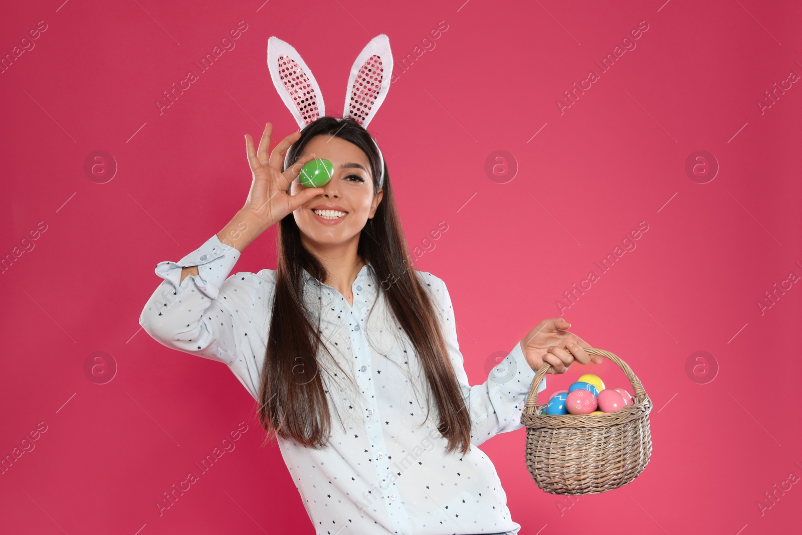 Photo of Beautiful woman in bunny ears headband with basket of Easter eggs on color background