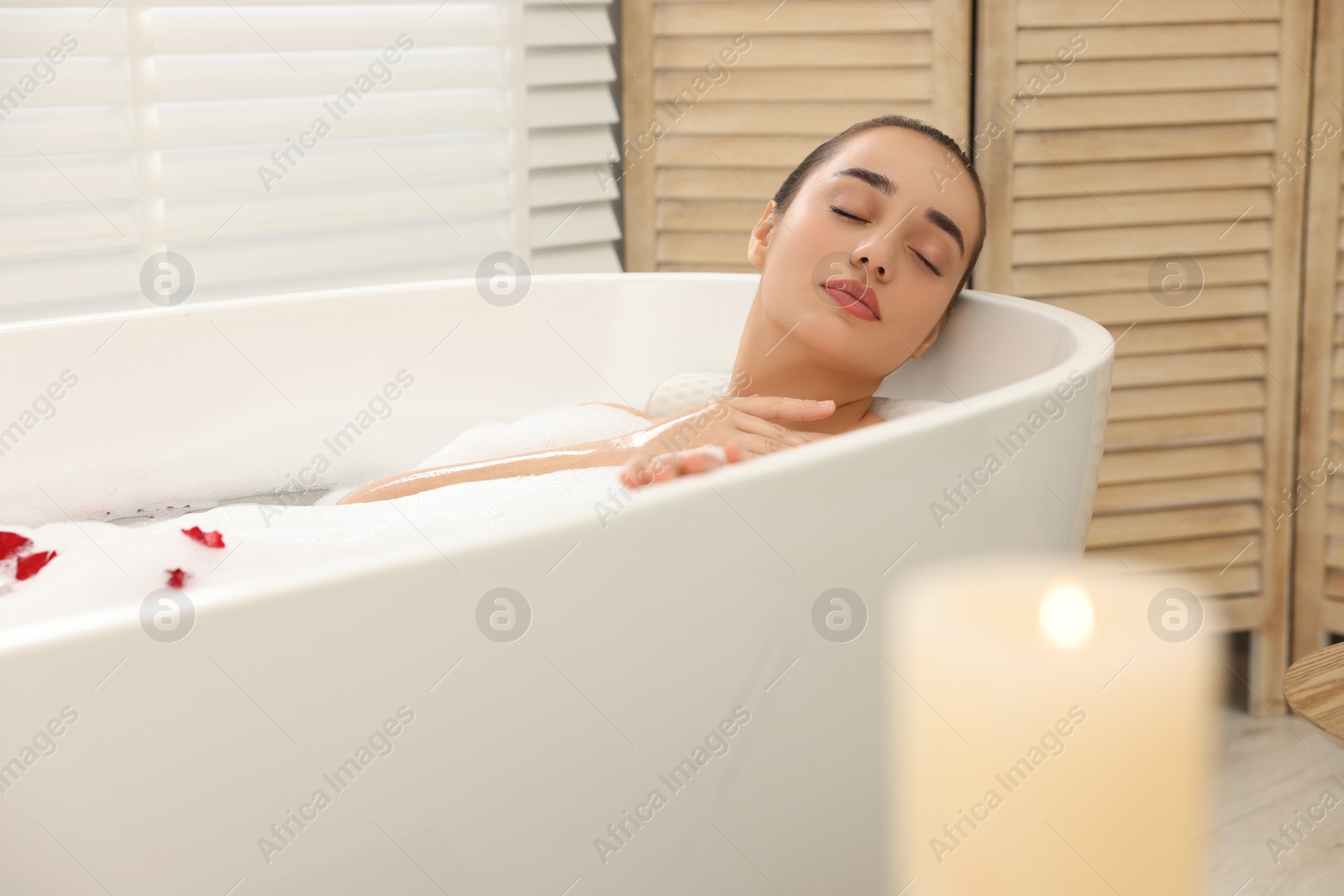 Photo of Woman taking bath in tub with foam and rose petals indoors