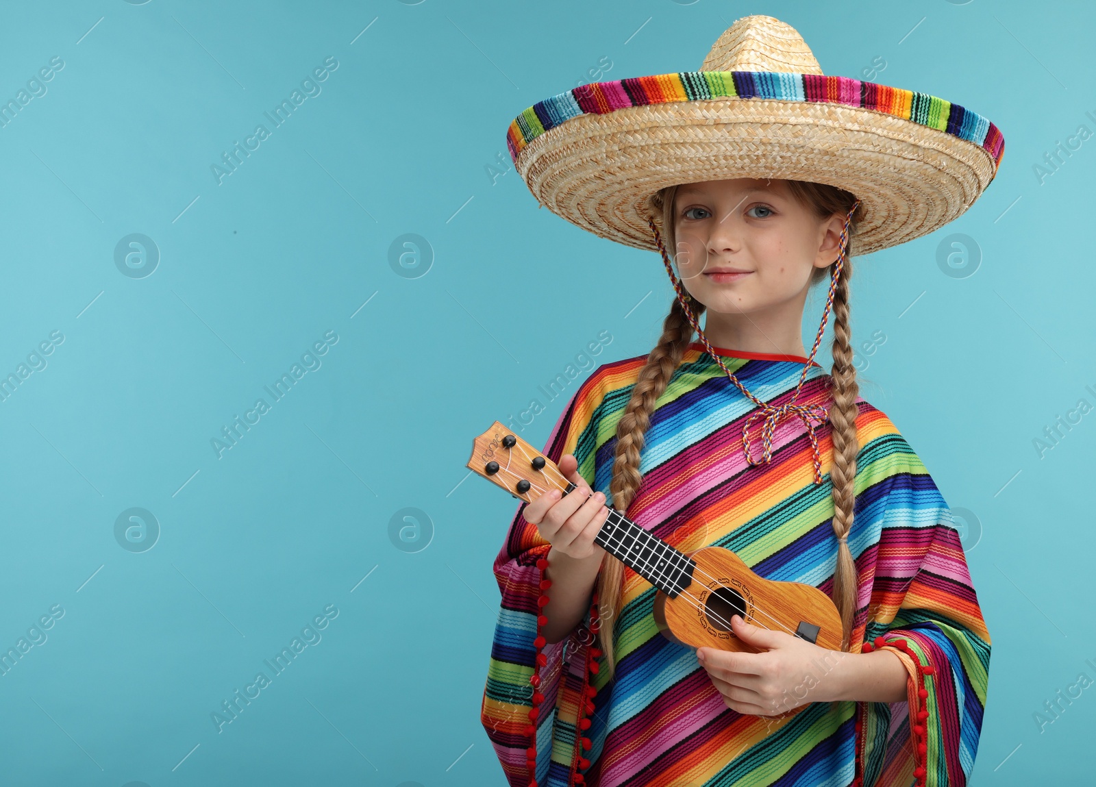 Photo of Cute girl in Mexican sombrero hat and poncho playing ukulele on light blue background. Space for text