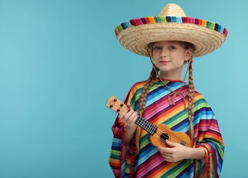 Cute girl in Mexican sombrero hat and poncho playing ukulele on light blue background. Space for text