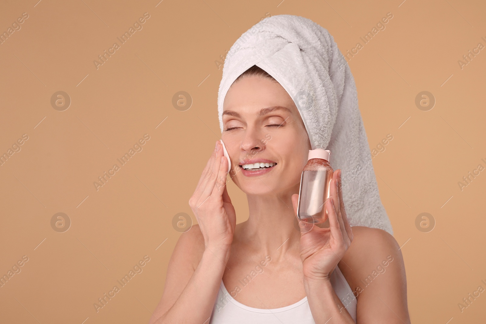 Photo of Beautiful woman in terry towel removing makeup with cotton pad on beige background, space for text
