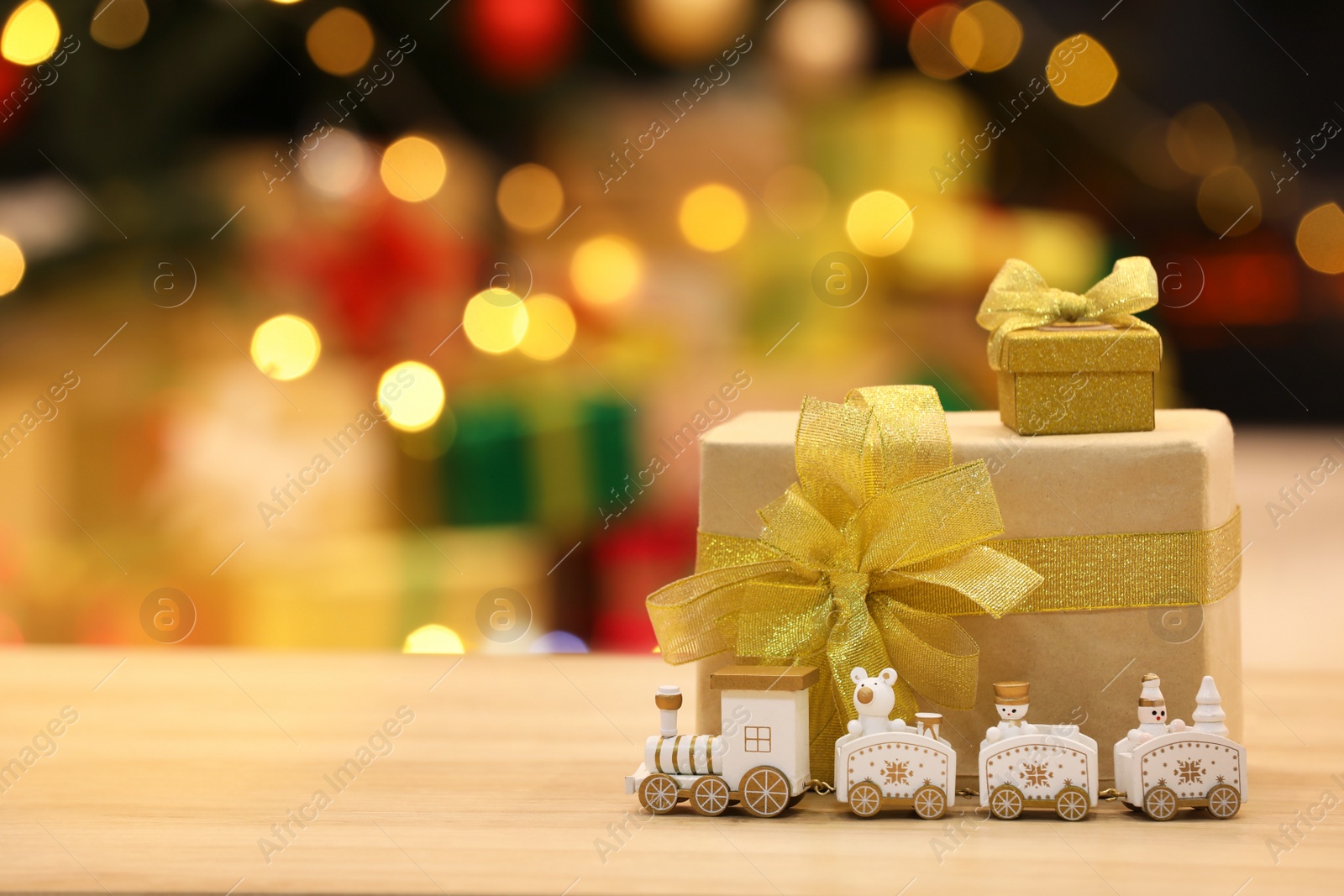 Photo of Beautiful gift boxes with toy train on wooden table against blurred festive lights, space for tex. Christmas celebration