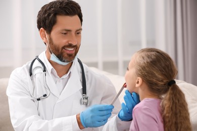 Smiling doctor examining girl`s oral cavity with tongue depressor indoors