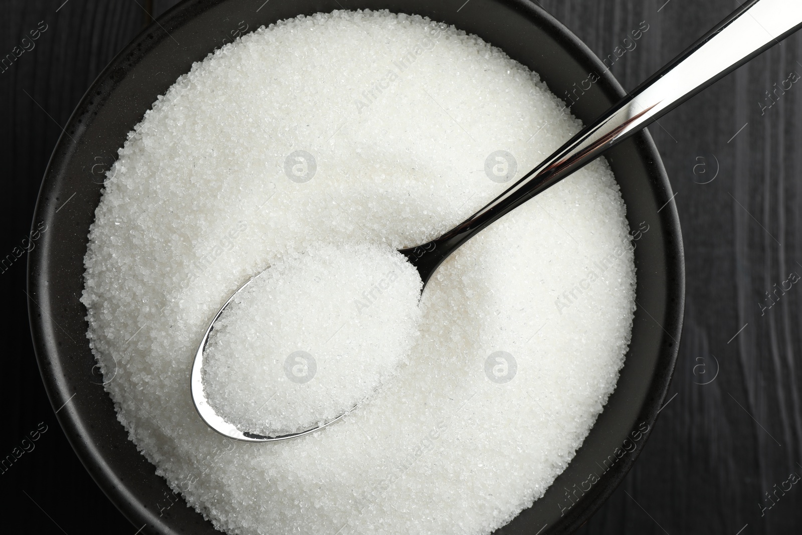 Photo of Granulated sugar in bowl and spoon on black wooden table, top view