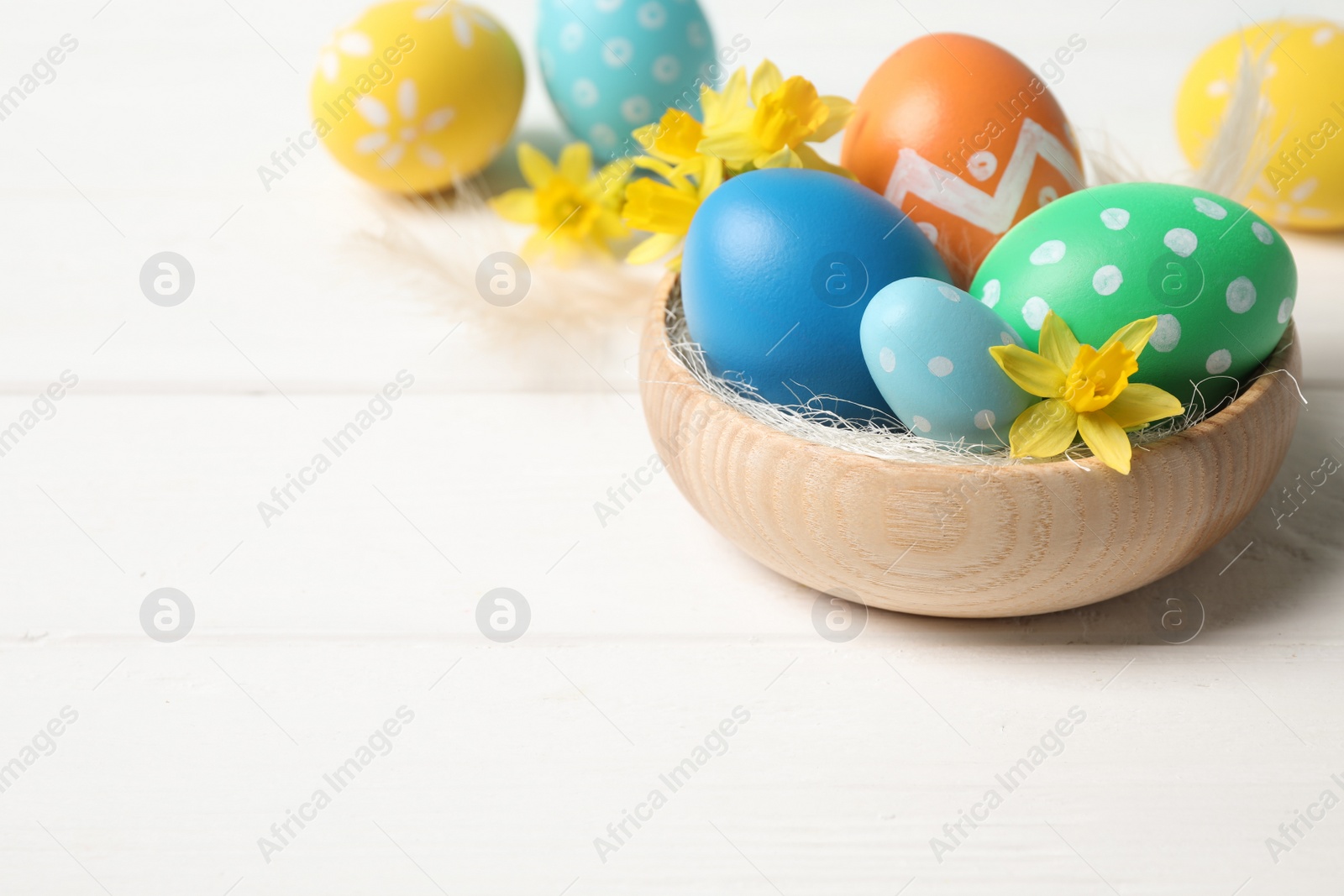 Photo of Colorful Easter eggs and flowers in bowl on white wooden table. Space for text